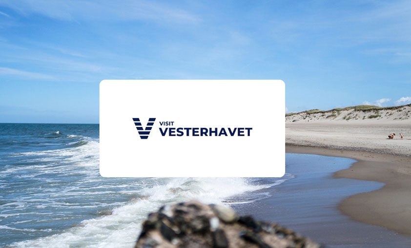 Visit Vesterhavets logo fremvises foran en idyllisk strandscene ved Vesterhavet, med blåt hav og sandstrand.