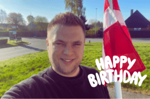 En mand smiler foran et dansk flag med teksten 'Happy Birthday' i hvid skrift.