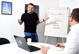 Mand i sort t-shirt præsenterer ved et whiteboard med teksten 'Valg af platform' for en anden person i hvid skjorte, der sidder ved et bord med en laptop og kaffekop.