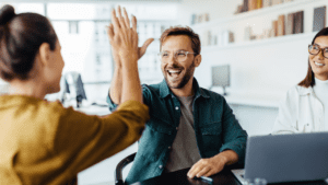 Smilende mand og kvindelig kollega i karrygul skjorte giver high-five på et kontor, med en asiatisk kvindelig kollega smilende ved siden af.